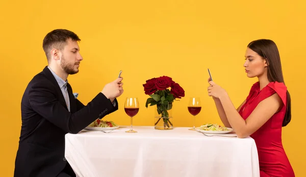 Casal usando celulares sentados na mesa posando sobre fundo amarelo — Fotografia de Stock