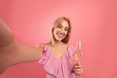 Şık giyinmiş gülümseyen genç bir kadın pembe stüdyo arka planında bir bardak şampanyayla selfie çekiyor.