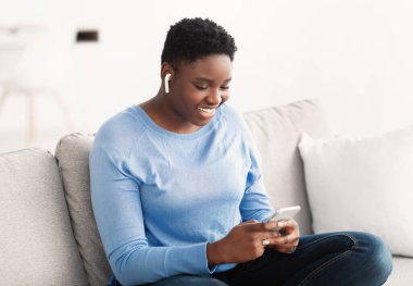 Gülümseyen siyah kadın akıllı telefon kullanıyor. Kulaklık takıyor.