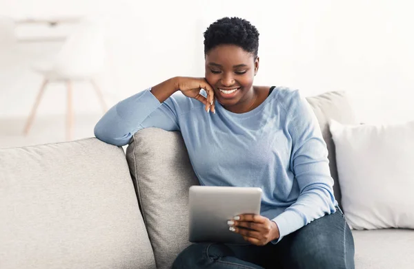 Siyah kadın kanepede oturuyor ve tablet kullanıyor. — Stok fotoğraf
