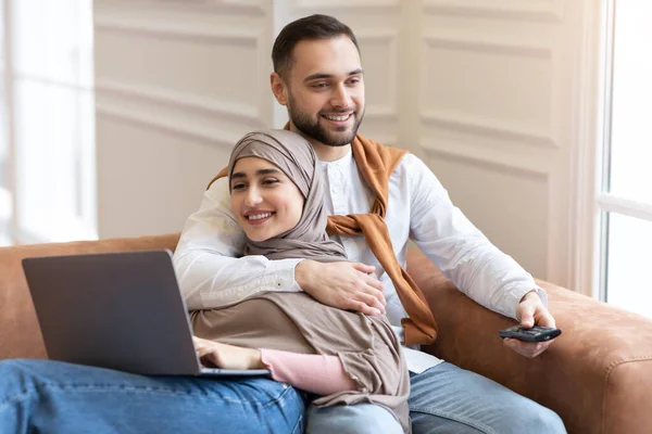 Мусульманская пара расслабляется с ноутбуком и смотрит телевизор дома — стоковое фото