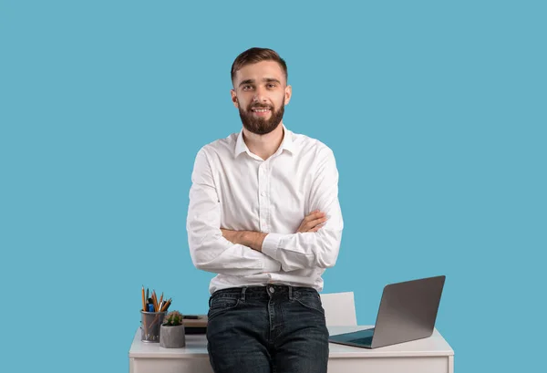 Jovem empresário feliz de pé com braços cruzados perto da mesa com computador portátil no fundo do estúdio azul — Fotografia de Stock