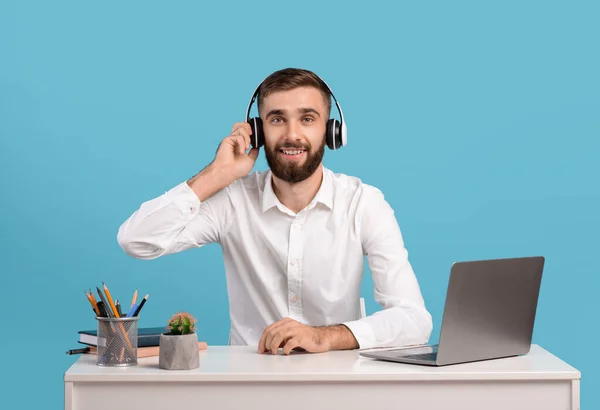 Operador de linha de apoio masculino barbudo com fones de ouvido e laptop respondendo chamada de clientes na mesa sobre fundo estúdio azul — Fotografia de Stock