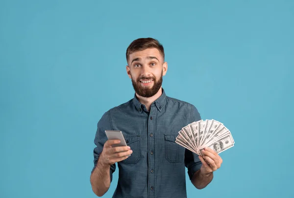 Crédito bancario en línea, lotería, casino. Feliz chico joven sosteniendo montón de dinero y teléfono inteligente en fondo de estudio azul —  Fotos de Stock