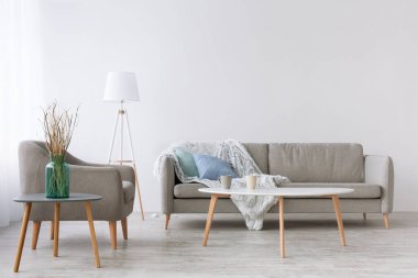 Mavi yastıklı kanepe ve yumuşak battaniye, koltuk, lamba ve iki fincanlı masa.