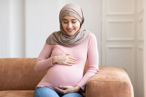 Embarazada musulmana dama en hiyab posando sentada en sofá interior — Foto de Stock