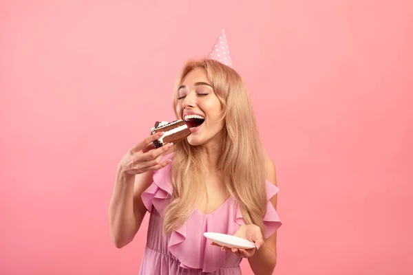 Mujer bastante joven comiendo delicioso pastel, con gorra de cumpleaños, celebrando las vacaciones en el fondo del estudio rosa — Foto de Stock