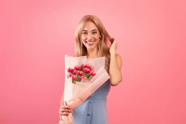 Gyönyörű hölgy szép ruha gazdaság tulipán, mosolygós és nézi a kamera felett rózsaszín stúdió háttér — Stock Fotó