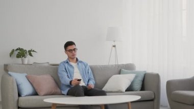 Gözlüklü genç adam evde televizyon izliyor, kanepede oturuyor ve kanal değiştiriyor.
