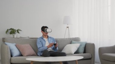 VR kulaklık takan, joystick ile video oyunu oynayan, evdeki kanepede oturan, ağır çekim