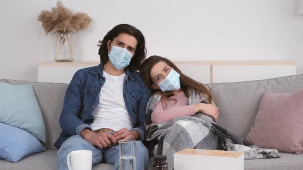 Coronavírus e auto-isolamento. Jovem casal deprimido vestindo máscaras de proteção médica sentado no sofá em casa — Vídeo de Stock