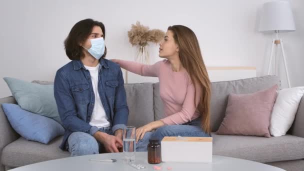 Jonge speelse dame wil haar vriendje kussen, maar de man met het medisch masker houdt haar tegen, zittend op de bank thuis — Stockvideo