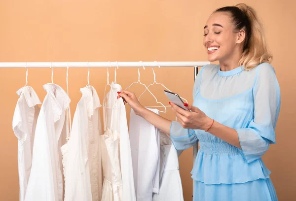 Wanita tersenyum memegang telepon berdiri di dekat rak dengan pakaian — Stok Foto