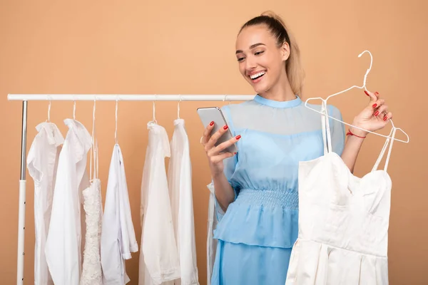 Usmívající se dáma pomocí telefonu stojí v blízkosti stojanu s oblečením — Stock fotografie