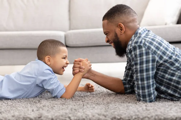 Afrykański ojciec i syn siłowanie się na rękę leży na podłodze w pomieszczeniach — Zdjęcie stockowe