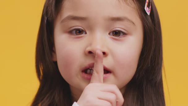 Тихо, это секрет. Крупным планом маленькая девочка кладет палец на губы, показывая жест молчания, оранжевый студийный фон — стоковое видео