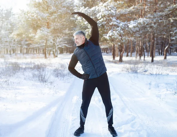 Deportes de temporada al aire libre. Retrato completo del chico maduro positivo haciendo ejercicios antes de trotar en el parque de invierno nevado —  Fotos de Stock