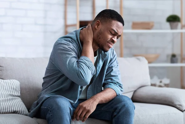 Hombre negro que sufre de dolor de cuello sentado en el sofá interior — Foto de Stock