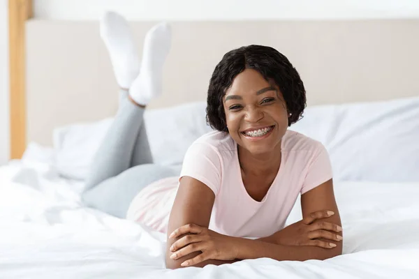 Положительная черная дама в пижаме лежит на кровати — стоковое фото