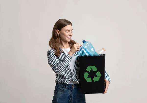 Переработка отходов. Улыбающаяся молодая женщина держит черный контейнер с пластиковыми бутылками — стоковое фото