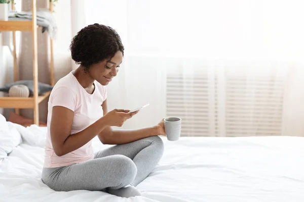 Sonriente dama africana en pijama bebiendo té, usando el teléfono — Foto de Stock