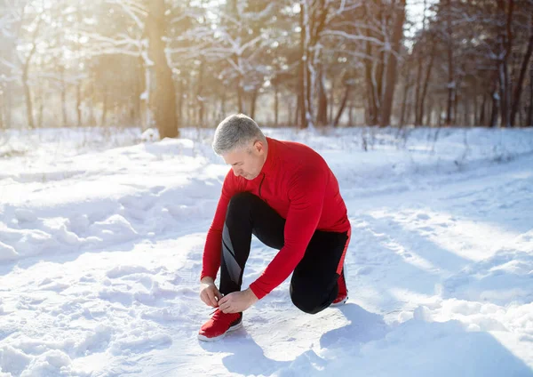 Mature jogger in warm sportswear tying shoelaces on snowy road at winter park — Stock Photo, Image