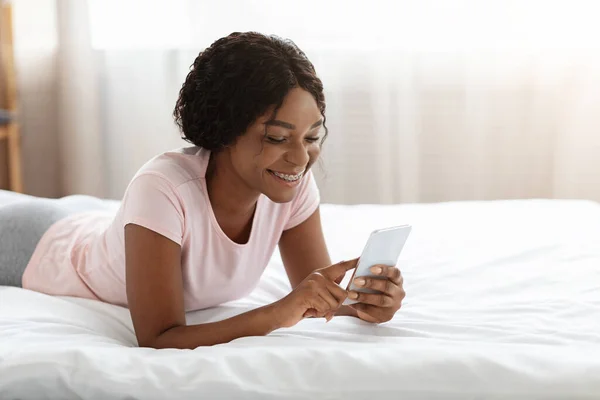 Feliz dama negra acostada en la cama, usando el teléfono móvil —  Fotos de Stock