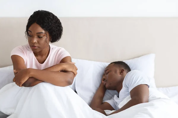 Upset african woman sitting in bed while her husband sleeping — Stock Fotó