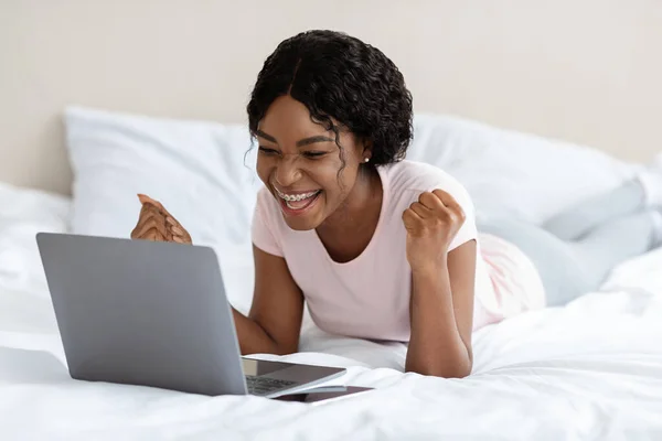Donna africana positiva che celebra il successo, posa con computer portatile sul letto — Foto Stock