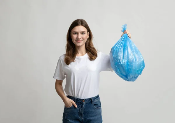 Wanita muda yang bahagia memegang kantong sampah plastik biru, berdiri di atas latar belakang abu-abu — Stok Foto