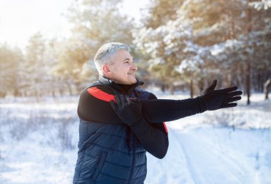 Winter outdoor workout concept. Cheerful mature guy doing warmup exercises before his run in snowy winter forest clipart