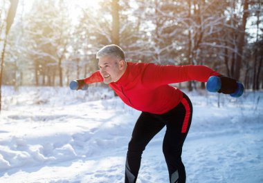 Açık hava kış dayanıklılık eğitimi. Dambılı yaşlı sporcu karlı parkta ağırlık kaldırma egzersizi yapıyor.