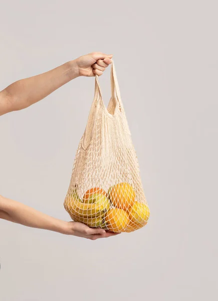 Manos sosteniendo la bolsa de malla ecológica con frutas orgánicas sobre fondo de estudio de luz —  Fotos de Stock