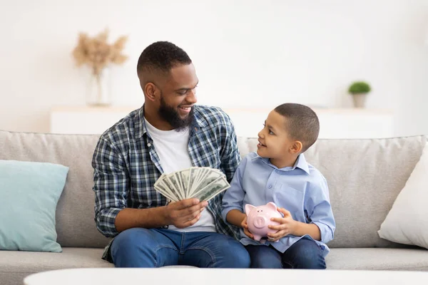 Black Father And His Son Putting Money In Piggybank Indoors