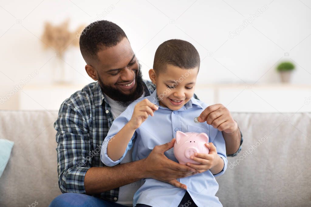 African Father And Son Putting Money In Piggybank At Home