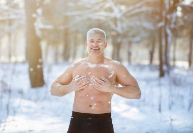 Cold exposure training concept. Joyful senior guy tempering his body with snow at frosty winter forest clipart