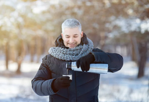 Hombre mayor feliz en chaqueta caliente y bufanda vertiendo té de termo frasco en el parque de invierno nevado. Actividades estacionales —  Fotos de Stock