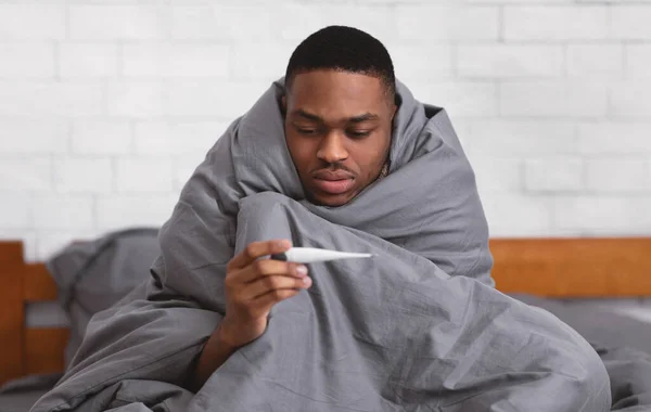 Hombre negro enfermo sosteniendo termómetro sentado envuelto en manta en interiores — Foto de Stock