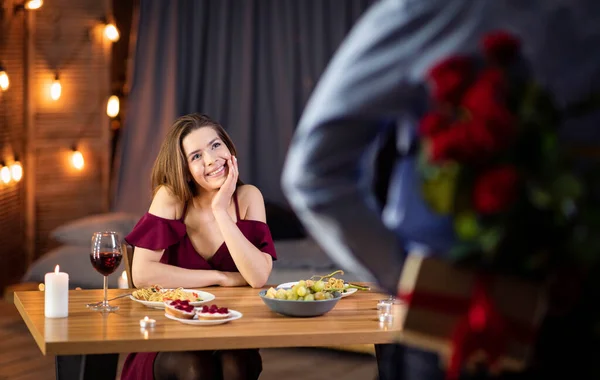Mężczyzna robi romantyczną niespodziankę dziewczynie w restauracji, trzymając róże i prezenty — Zdjęcie stockowe