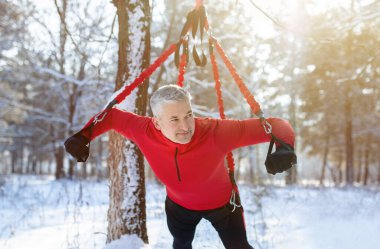 Outdoor bungee fitness. Athletic senior man working out with suspension training straps at snowy winter forest clipart