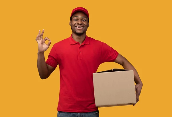 Czarny dostawca trzyma tekturowe pudełko z dobrym gestem. — Zdjęcie stockowe