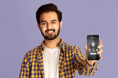 Violet stüdyosunun arka planında cep telefonu ve taksi hizmeti uygulaması gösteren pozitif Hintli adam.