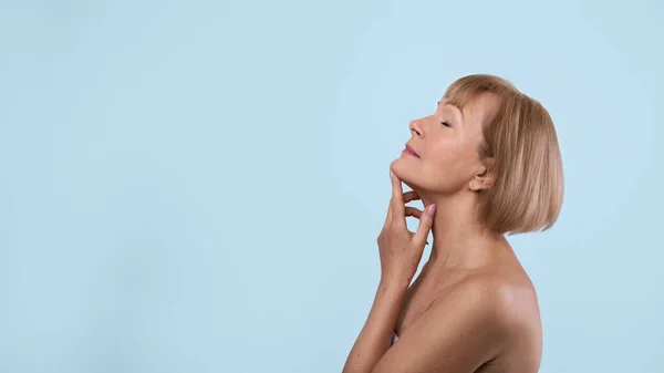 Grazioso invecchiamento. Bella signora anziana toccare la pelle sul collo su sfondo blu. Panorama vista laterale con spazio copia — Foto Stock