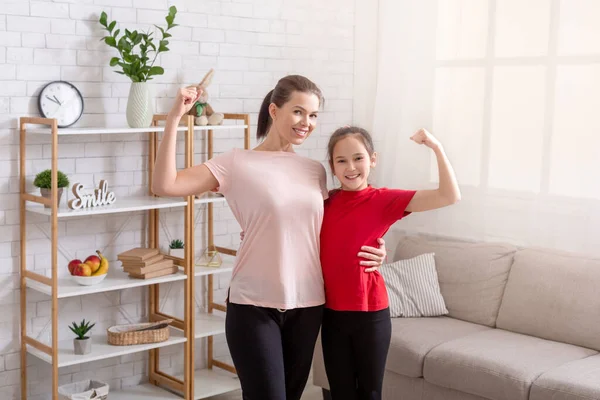 Портрет улыбающейся матери и дочери-подростка в спортивной одежде, показывающей свои бицепсные мышцы, демонстрирующей силу дома — стоковое фото