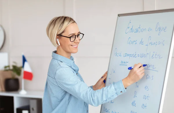 Vriendelijke vrouwelijke leraar die grammatica regels schrijft op schoolbord tijdens online Franse les. Onderwijs en bijscholing op afstand — Stockfoto