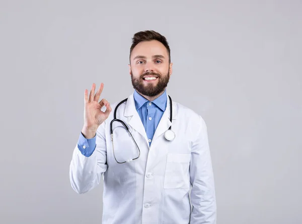 Portrait of happy young doctor in uniform showing okay gesture on grey studio background. Healthcare concept — Stock Photo, Image