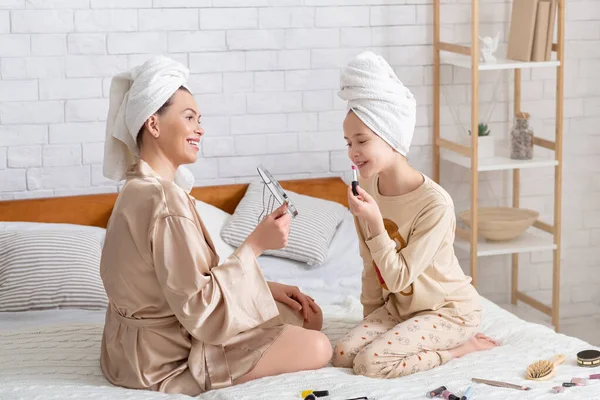Mujer encantadora y su hija en ropa de casa y toallas de baño que se divierten con el maquillaje, la aplicación de lápiz labial en la cama en casa —  Fotos de Stock