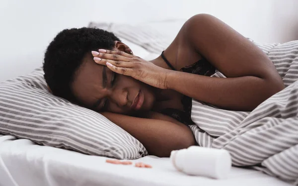 Молодая африканская дама с закрытыми глазами прижимает руки к искушениям и задыхается от головной боли, лежащей на кровати — стоковое фото