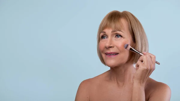 Naturlig makeup för äldre kvinnor. Porträtt av vackra senior kvinna tillämpa kosmetika på hennes hud över blå bakgrund — Stockfoto