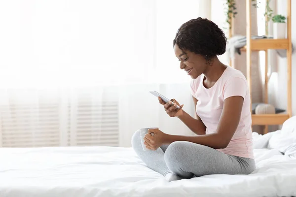 Mujer negra alegre sentada en la cama, usando el teléfono móvil — Foto de Stock